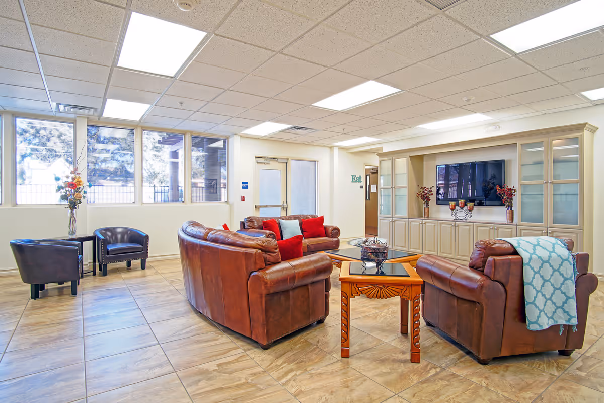 A bright and spacious living room area with large windows letting in natural light. The room features brown leather sofas with red and blue cushions, a wooden coffee table, and a side table with a decorative vase of flowers. There is a large built-in entertainment center with a flat-screen TV and decorative items. The floor is tiled, and the ceiling has recessed lighting panels.
