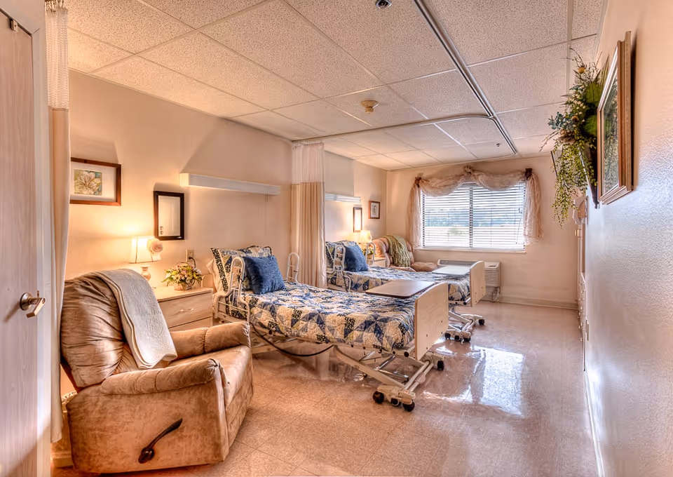 A well-lit room in a senior living facility with two hospital-style beds covered in blue and white patchwork quilts, each with a blue pillow. There is a beige recliner chair with a white blanket draped over it, a small wooden nightstand with a lamp and flowers, and a window with blinds and sheer curtains letting in natural light. The walls are decorated with framed pictures and a hanging plant.