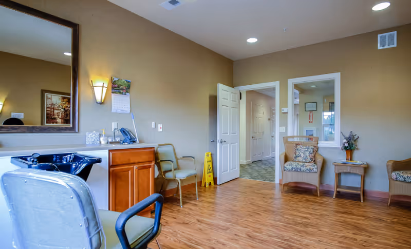 A clean and well-lit room with wooden flooring, beige walls, and a large mirror above a countertop with a black sink. There are several chairs, including a salon chair and cushioned armchairs, a small table with a flower vase, and an open door leading to a hallway. A yellow caution sign is visible near the door.