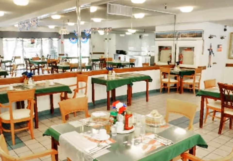 A dining room with multiple tables covered in green tablecloths and set with plates, cups, and condiments. Wooden chairs surround the tables, and the room features tiled flooring and large mirrors on one wall. Decorations hang from the ceiling, and there are windows letting in natural light.