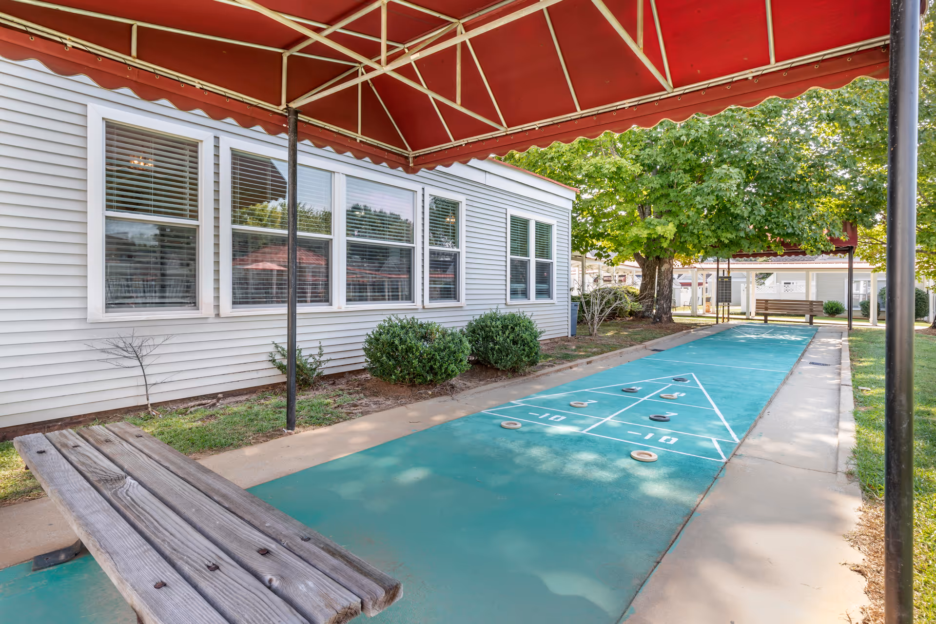 Outdoor shuffleboard court with a green playing surface and several shuffleboard discs scattered on it, covered by a red canopy. There is a wooden bench in the foreground and a large tree with green leaves in the background near a white building with multiple windows.