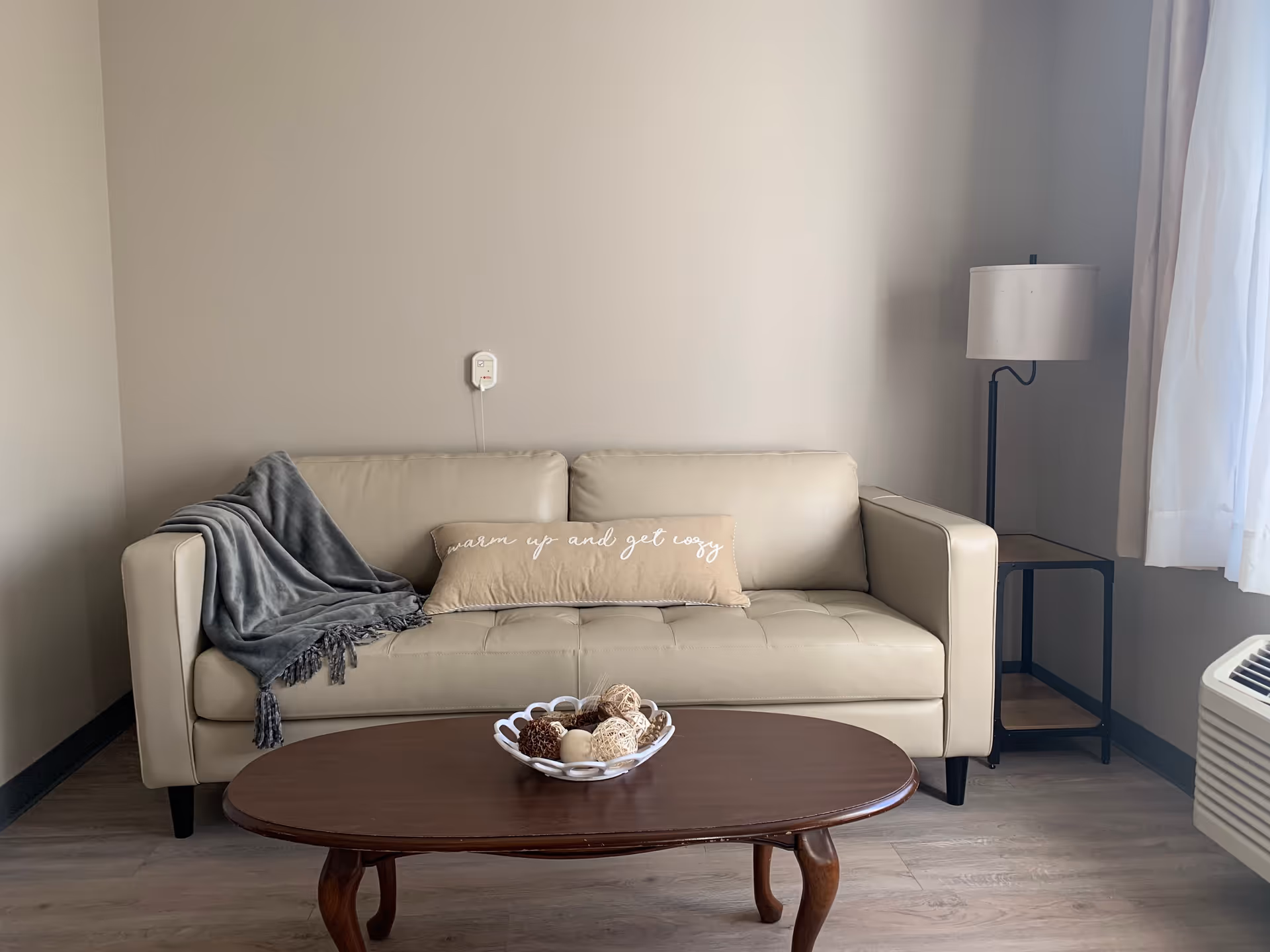 A beige leather sofa with a gray throw blanket on the left armrest and a beige rectangular pillow with the text 'warm up and get cozy' sits against a plain light-colored wall. In front of the sofa is a wooden coffee table with a decorative bowl containing various natural elements. To the right of the sofa is a floor lamp with a white lampshade and a small side table. A window with white curtains and an air conditioning unit are visible on the right side of the image.