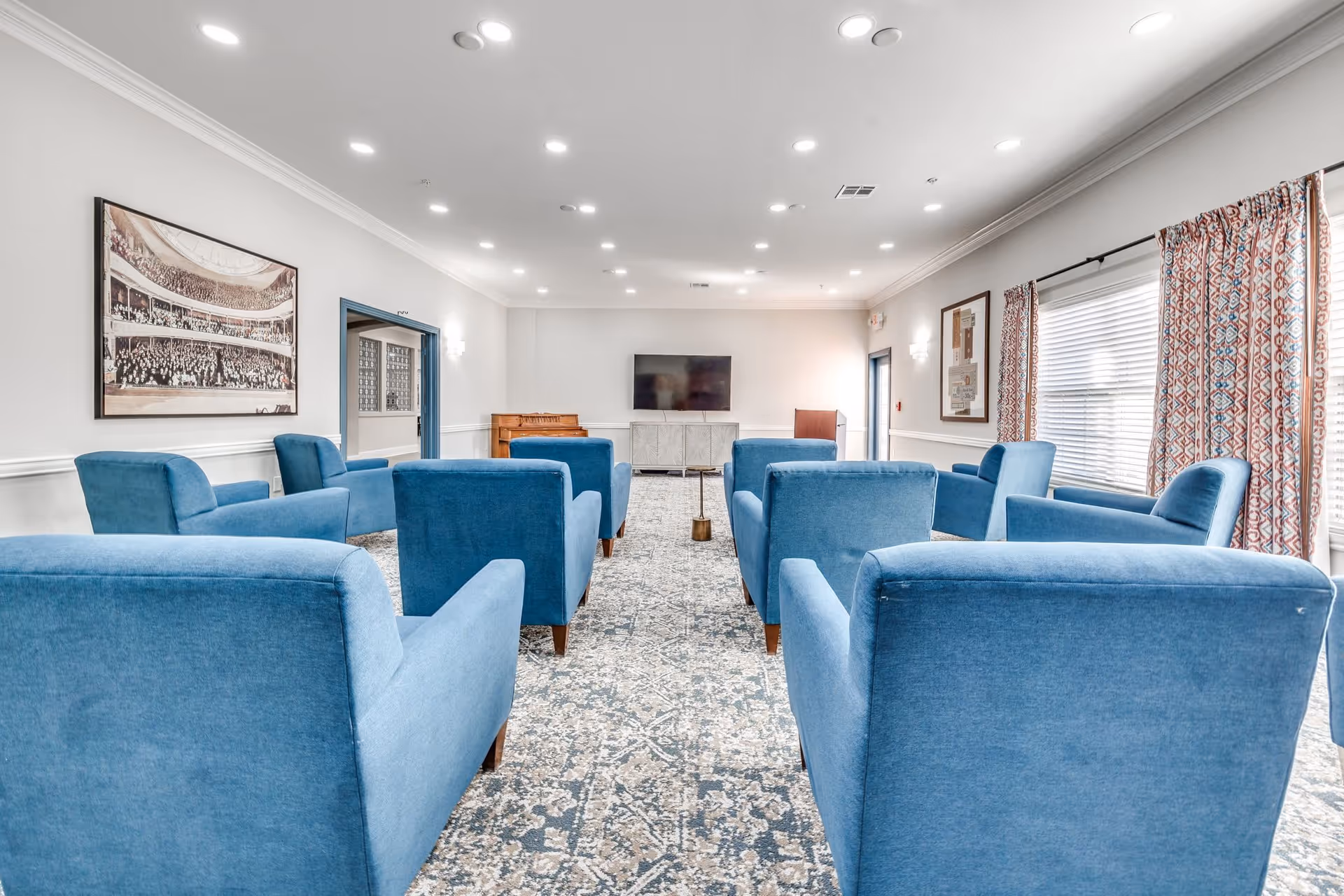 A senior living facility common room with multiple blue upholstered armchairs arranged in rows facing a wall-mounted flat screen TV. The room has a patterned carpet, white walls with framed artwork, large windows with patterned curtains, and recessed ceiling lights.