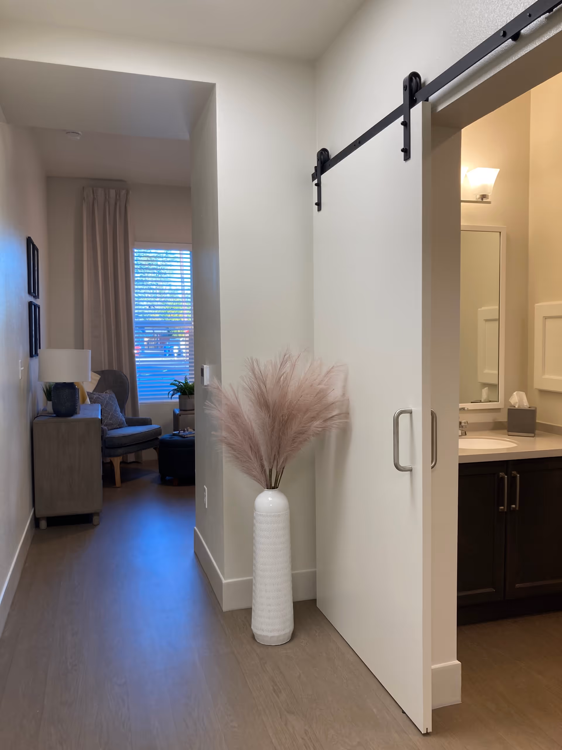 Interior view of a hallway in an assisted living facility with a white sliding barn door partially open, revealing a bathroom sink and mirror. A tall white vase with decorative pampas grass is placed against the wall. In the background, there is a cozy sitting area with a chair, side table, lamp, and window with blinds and curtains.