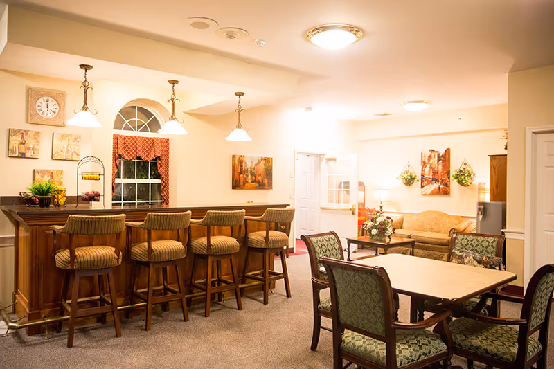 A cozy interior common area featuring a wooden bar with five cushioned bar stools, a table with four upholstered chairs, and a sitting area with a sofa and armchairs. The room is warmly lit with ceiling lights and pendant lamps, decorated with framed artwork, plants, and a clock above the bar.