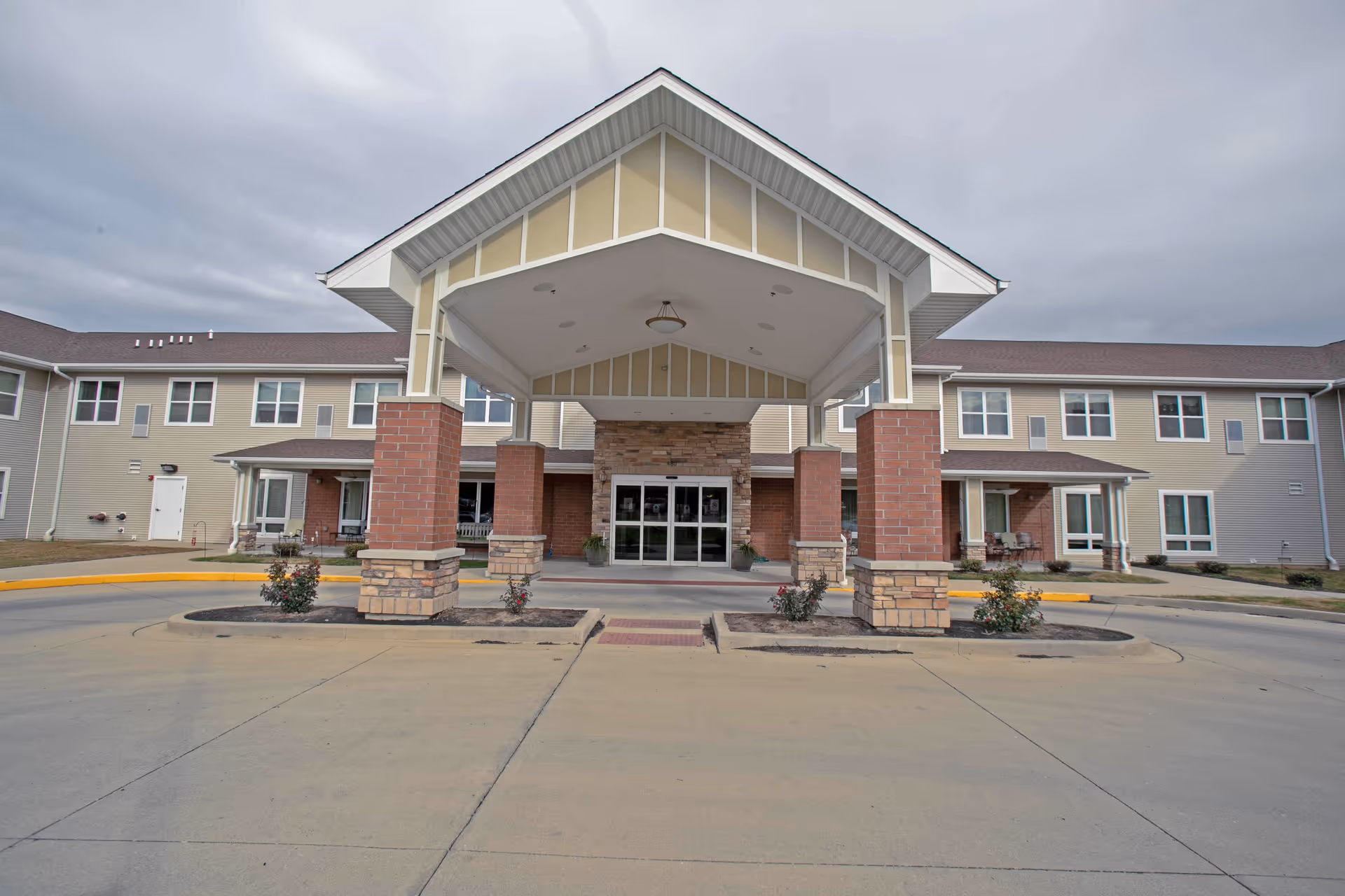 Front exterior view of a two-story senior living facility building with a covered entrance supported by brick and stone columns. The building has multiple windows and a driveway leading up to the entrance under a cloudy sky.