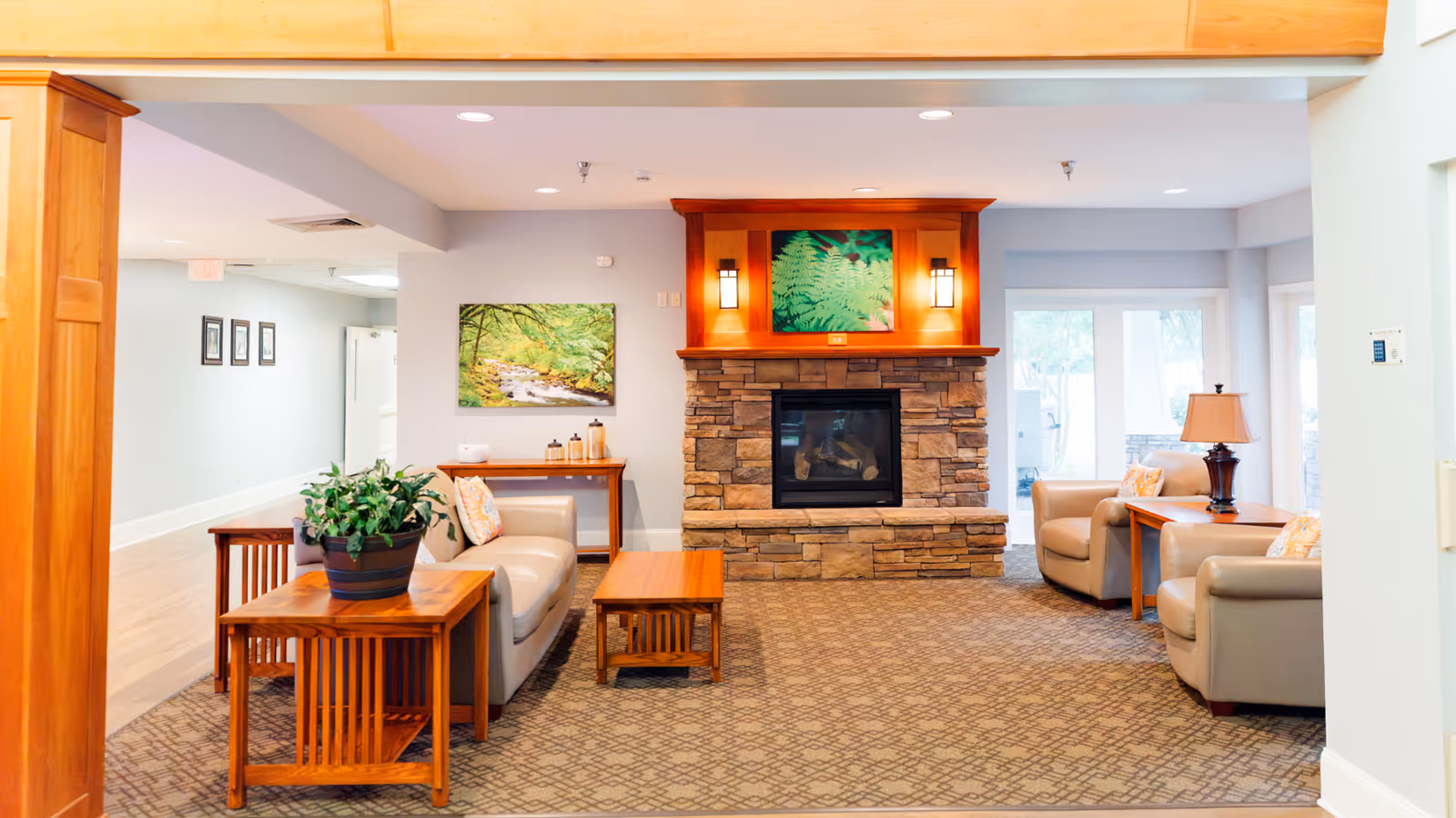 A cozy living room area with a stone fireplace in the center, flanked by two wall-mounted lights and a framed picture above. The room features beige leather armchairs and a sofa, wooden side tables with a potted plant and a lamp, and a patterned carpet. Large windows and glass doors allow natural light to enter the space.