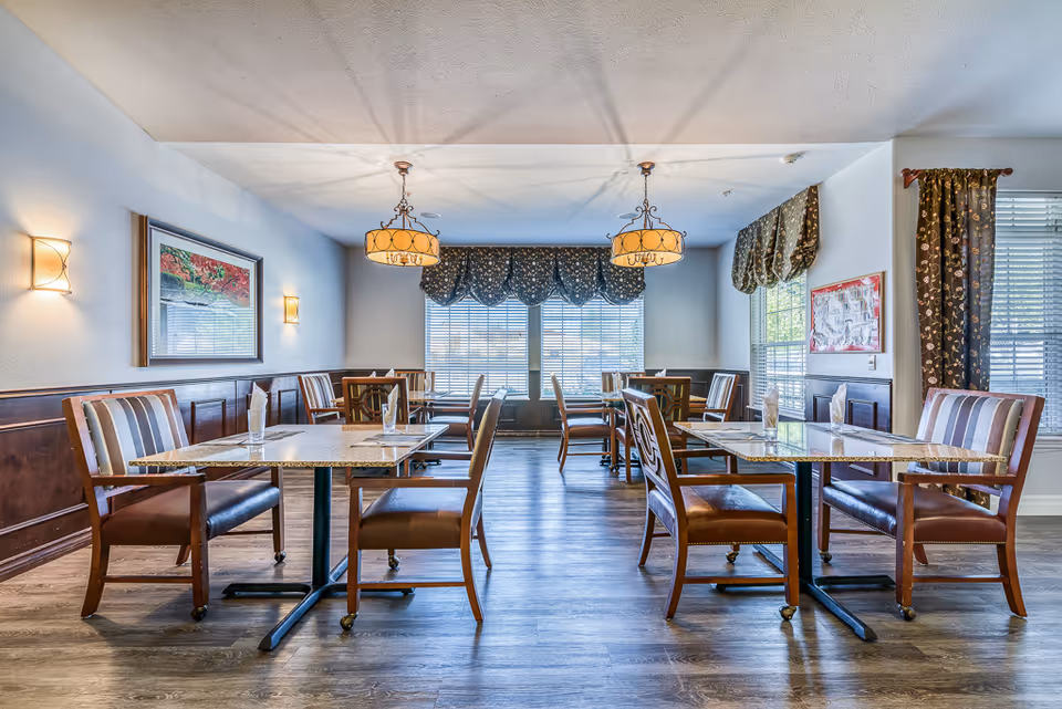 A bright dining room in Dalton Senior Living with multiple tables and chairs arranged neatly. The room features wooden flooring, large windows with patterned valances and curtains, wall sconces, and two hanging pendant lights. Each table has a glass and a folded napkin placed on it.