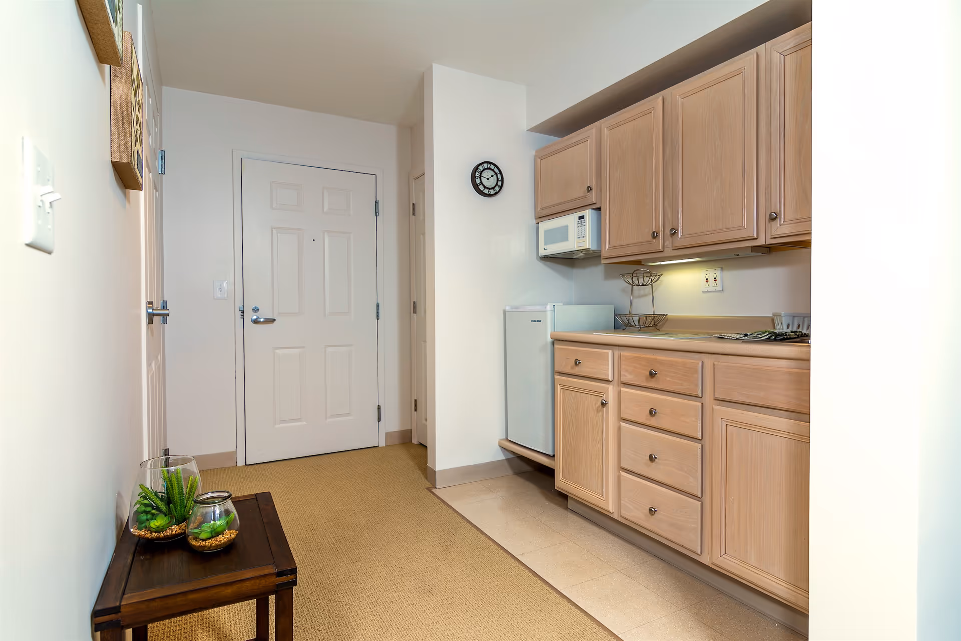 Interior view of a small kitchen area in a memory care facility with light wood cabinets, a microwave, a mini refrigerator, and a countertop with a two-tier fruit basket. A small wooden table with two glass containers holding green plants is placed against the opposite wall. A white door and a clock are visible in the background.