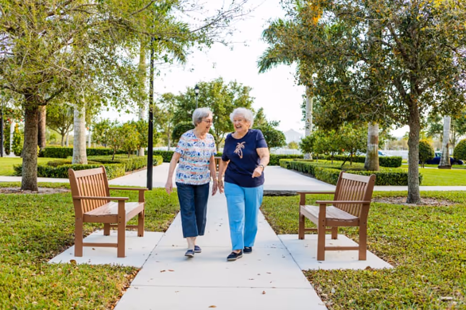 Two people walking along a tree-lined sidewalk between wooden benches in a landscaped outdoor area.