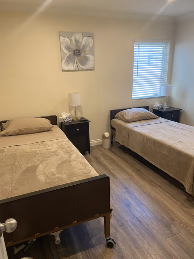 A simple bedroom with two single beds, each with beige bedding and pillows. Between the beds are two black nightstands, each with a white lamp and some tissues. A window with white blinds is on the right wall, and a large flower painting hangs on the wall above the left nightstand. The floor is wood laminate.