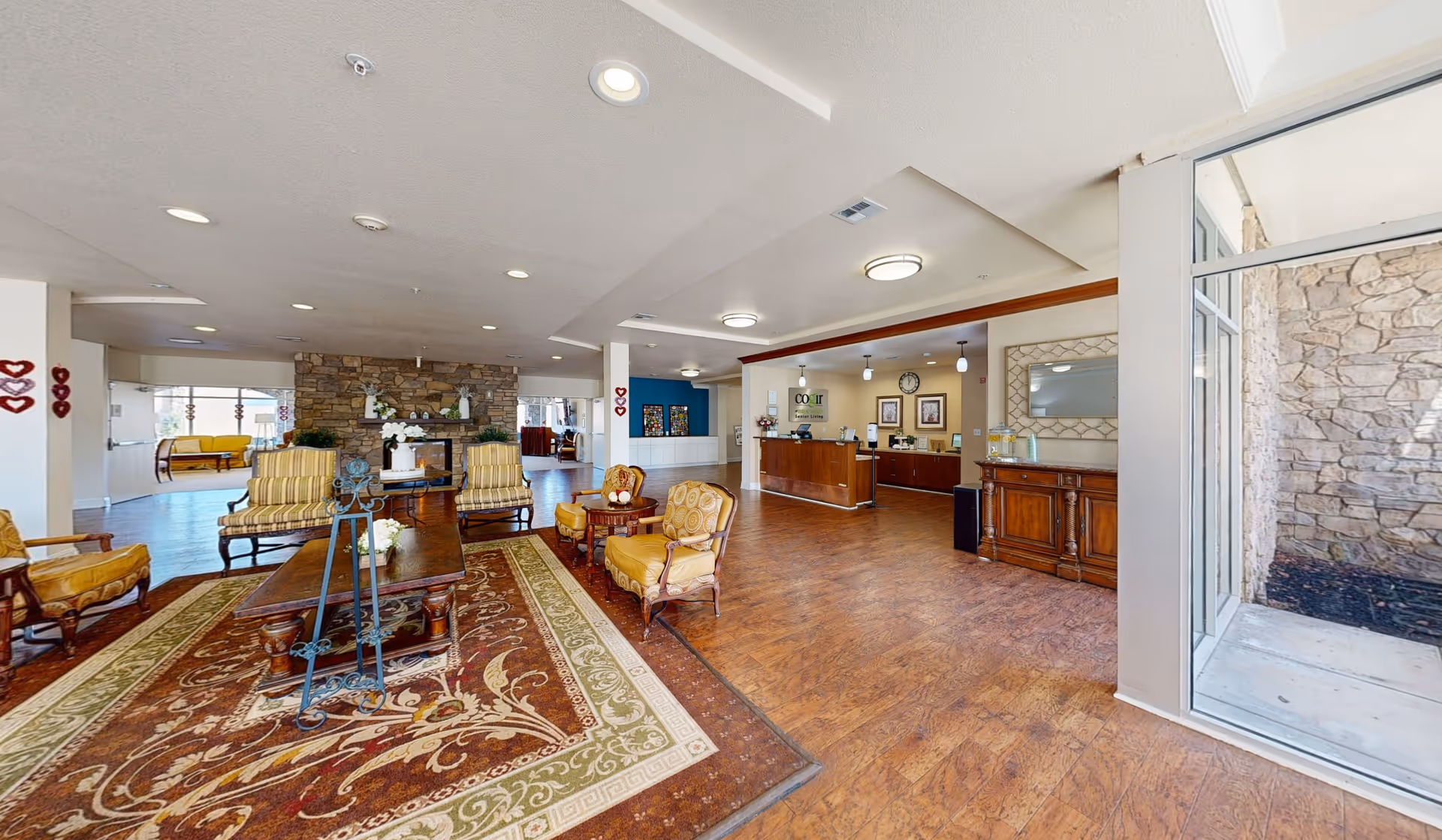 Spacious senior living facility lobby with wooden flooring, a large patterned area rug, several upholstered armchairs and a wooden coffee table. A stone fireplace is visible in the background along with a reception desk and decorative wall art. Large windows allow natural light to enter the space.