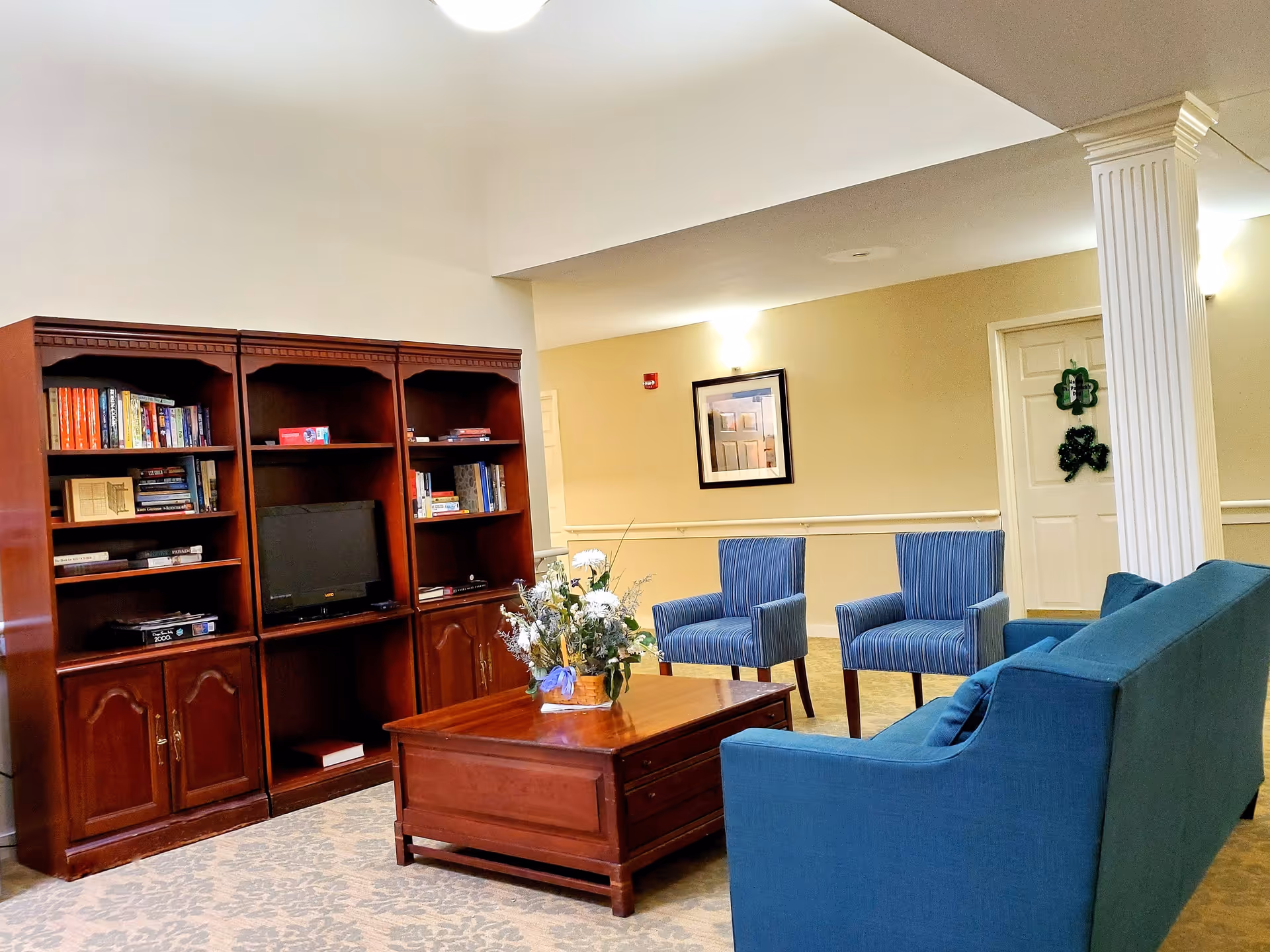 A cozy living room area in a senior living facility featuring a wooden entertainment center with books and a TV, a wooden coffee table with a floral arrangement, two blue striped armchairs, and a blue sofa. The room has beige walls, a framed picture, and a white column near the seating area.