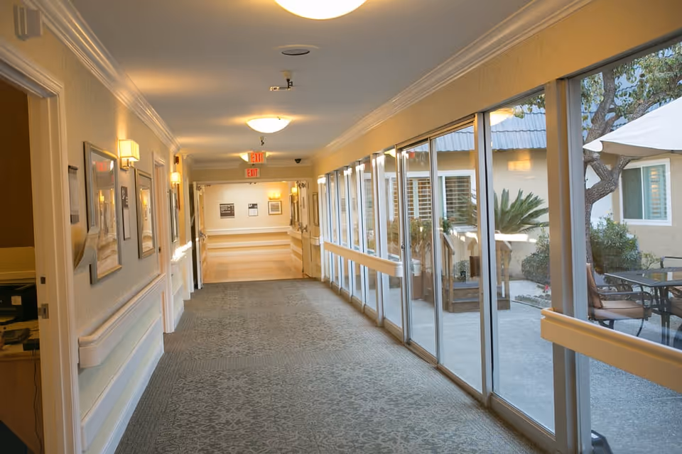 A well-lit indoor hallway in a senior living facility with carpeted floor and handrails along the walls. On the right side, large glass windows and sliding doors reveal an outdoor patio area with tables, chairs, plants, and an umbrella. The hallway has framed pictures on the left wall and exit signs visible at the far end.