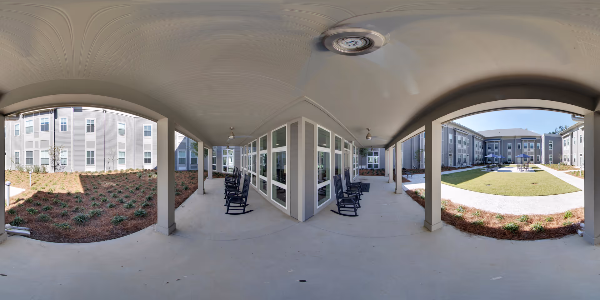Covered outdoor patio area with ceiling fans and several black rocking chairs facing a landscaped courtyard with grass, pathways, and surrounding multi-story buildings under a clear blue sky.