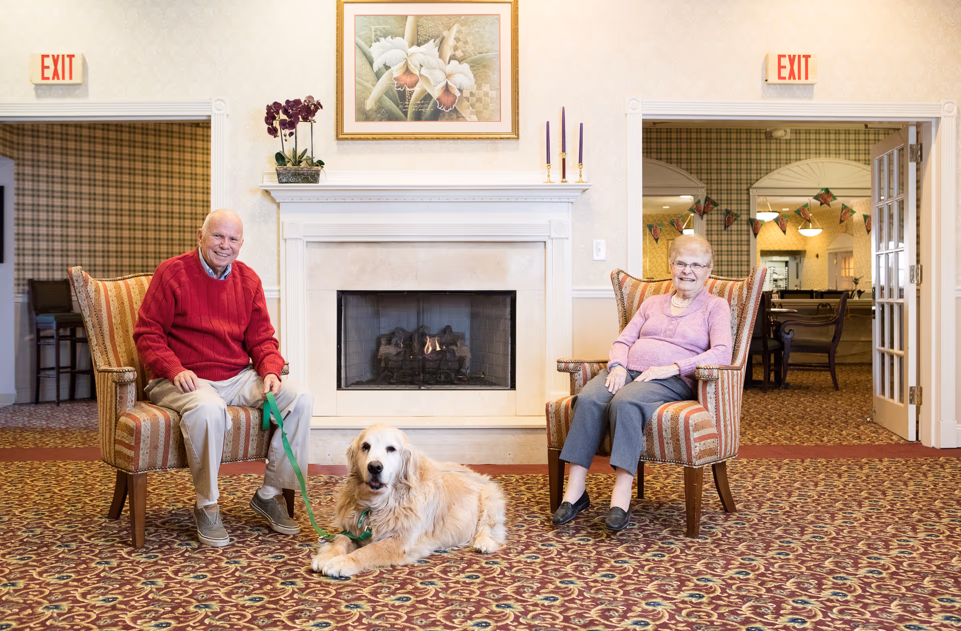 An elderly man and woman sitting in striped armchairs on either side of a fireplace in a cozy room. The man is holding a leash attached to a golden retriever lying on a patterned carpet in front of them. There are exit signs above doorways on both sides of the fireplace, and a framed floral painting hangs above the mantel.