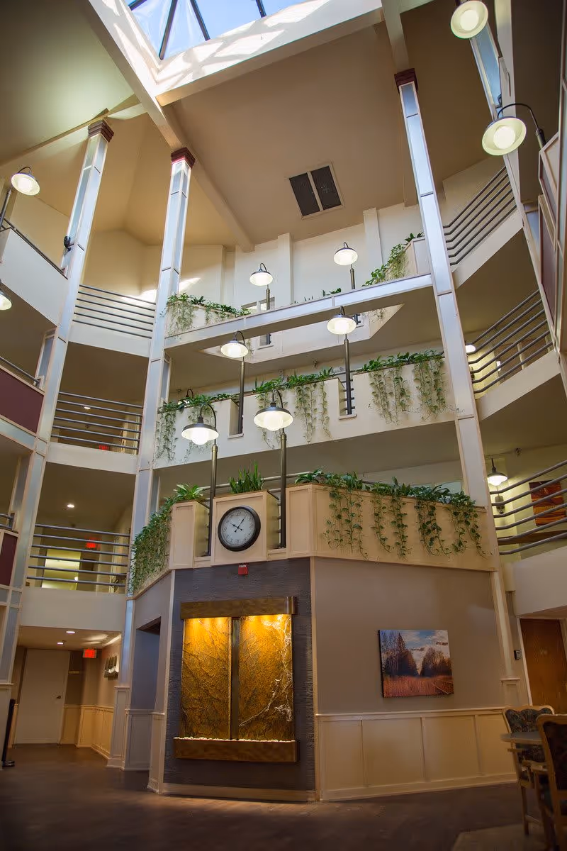 Interior view of a multi-story atrium in a senior living facility with hanging lamps, green plants cascading from railings, a large clock mounted above a decorative water feature, and a skylight ceiling allowing natural light to enter.