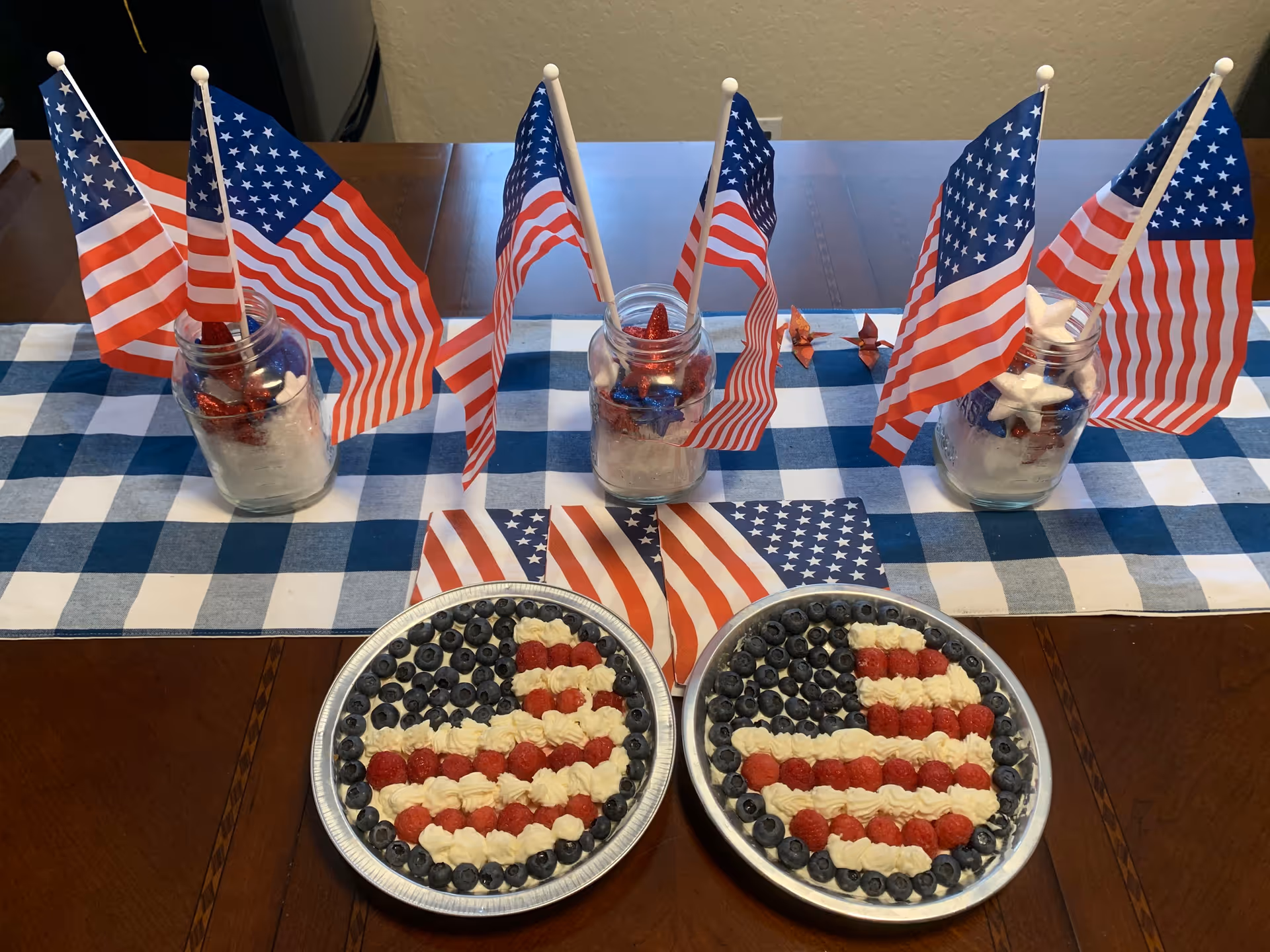 Two round fruit tarts decorated as American flags on a table with a blue-and-white checkered runner and mason jars holding small U.S. flags.