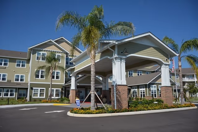Exterior view of Orchard Heights Gracious Retirement Living showing a multi-story building with green siding and white trim, palm trees, and a covered entrance with brick pillars. The driveway and landscaped areas with flowers are visible in the foreground under a clear blue sky.