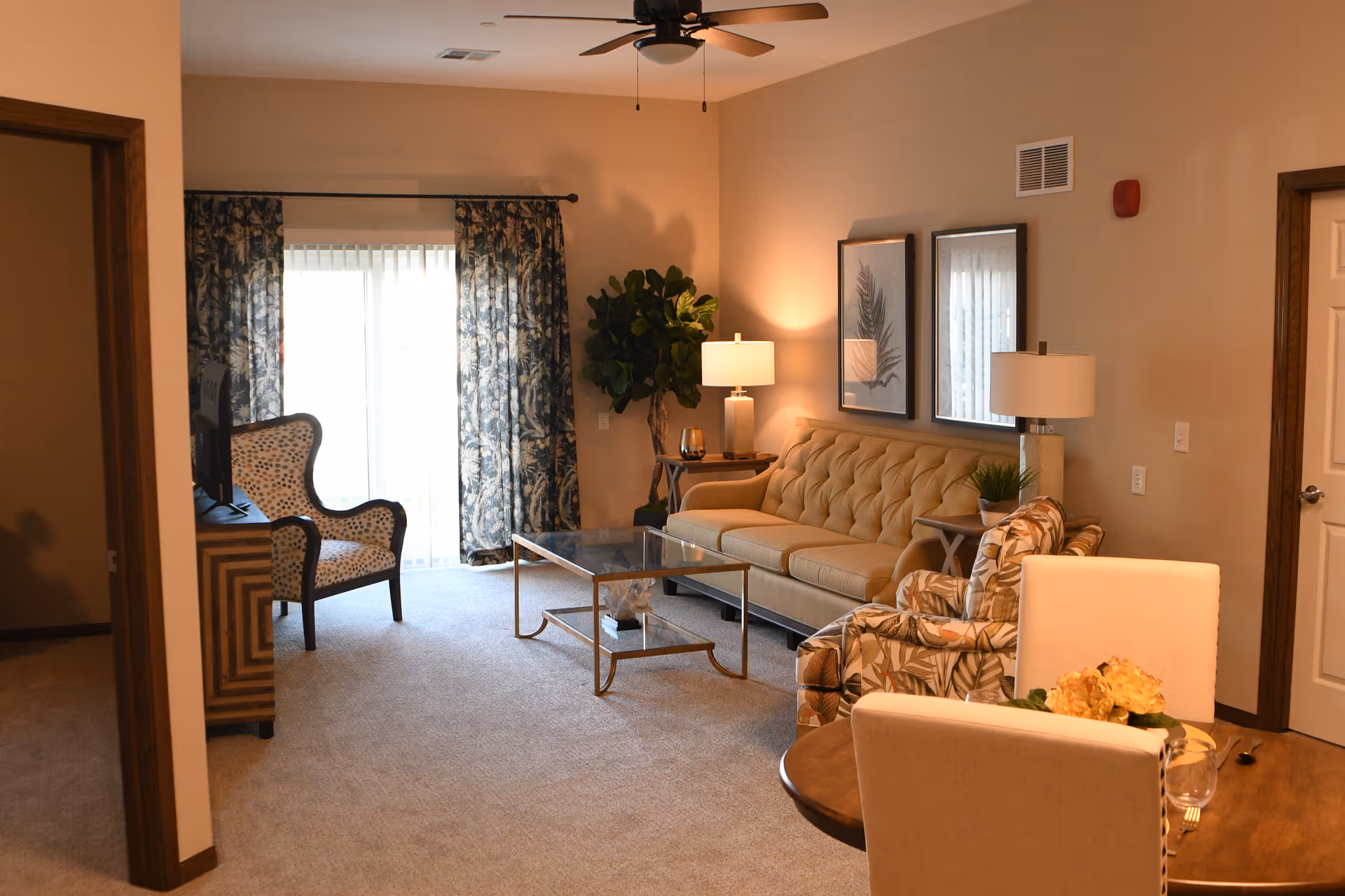 A cozy living room in Shell Harbor Retirement Community featuring a beige tufted sofa, a patterned armchair, a glass coffee table, two side tables with lamps, a ceiling fan, and a sliding glass door with floral curtains letting in natural light. There is also a small dining table with two chairs and a decorative plant in the corner.