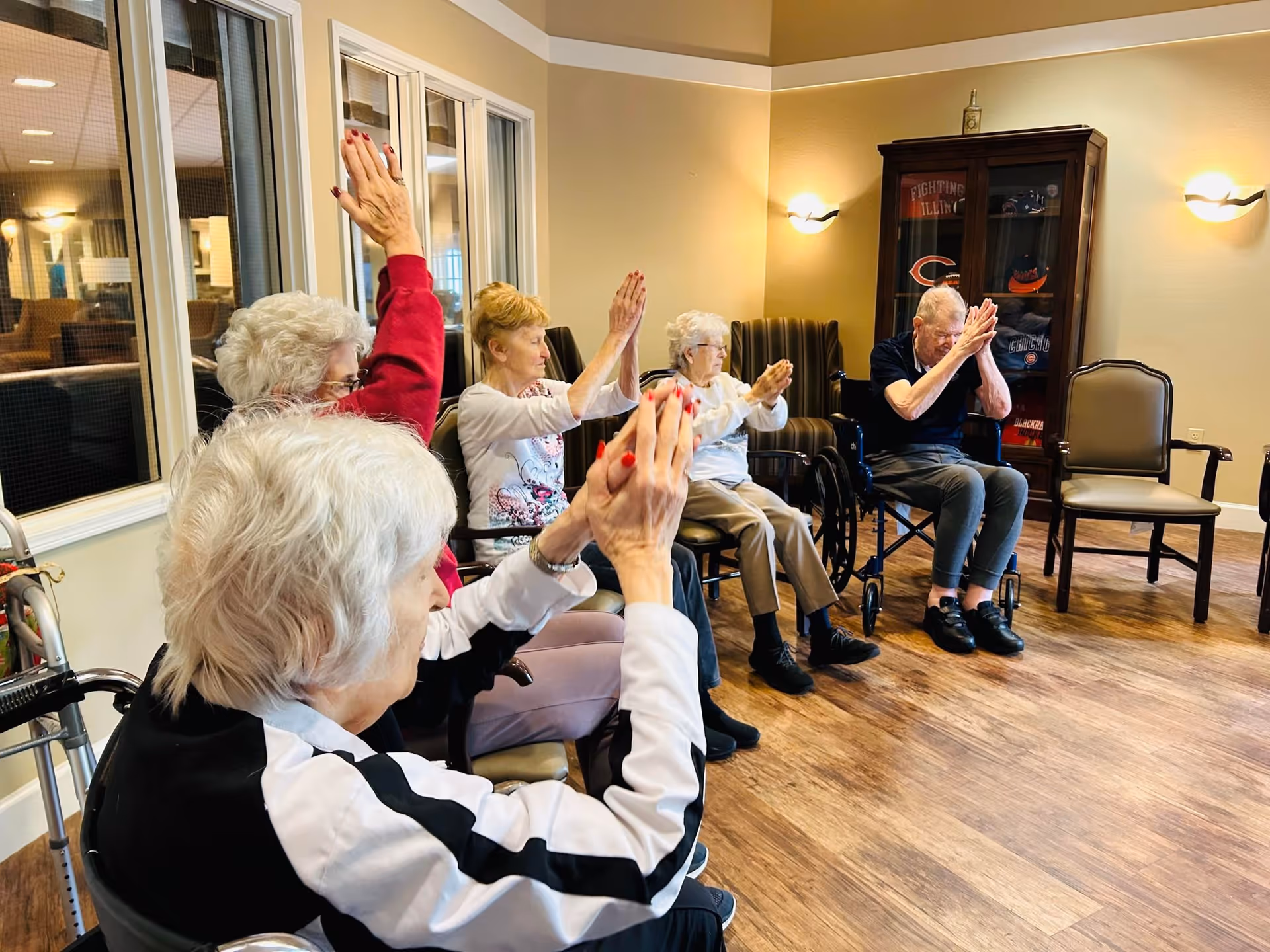 A group of elderly individuals seated in a circle in a well-lit room, participating in a seated exercise or activity with their hands raised. Some are in wheelchairs and others in chairs, with a wooden cabinet and wall lights in the background.