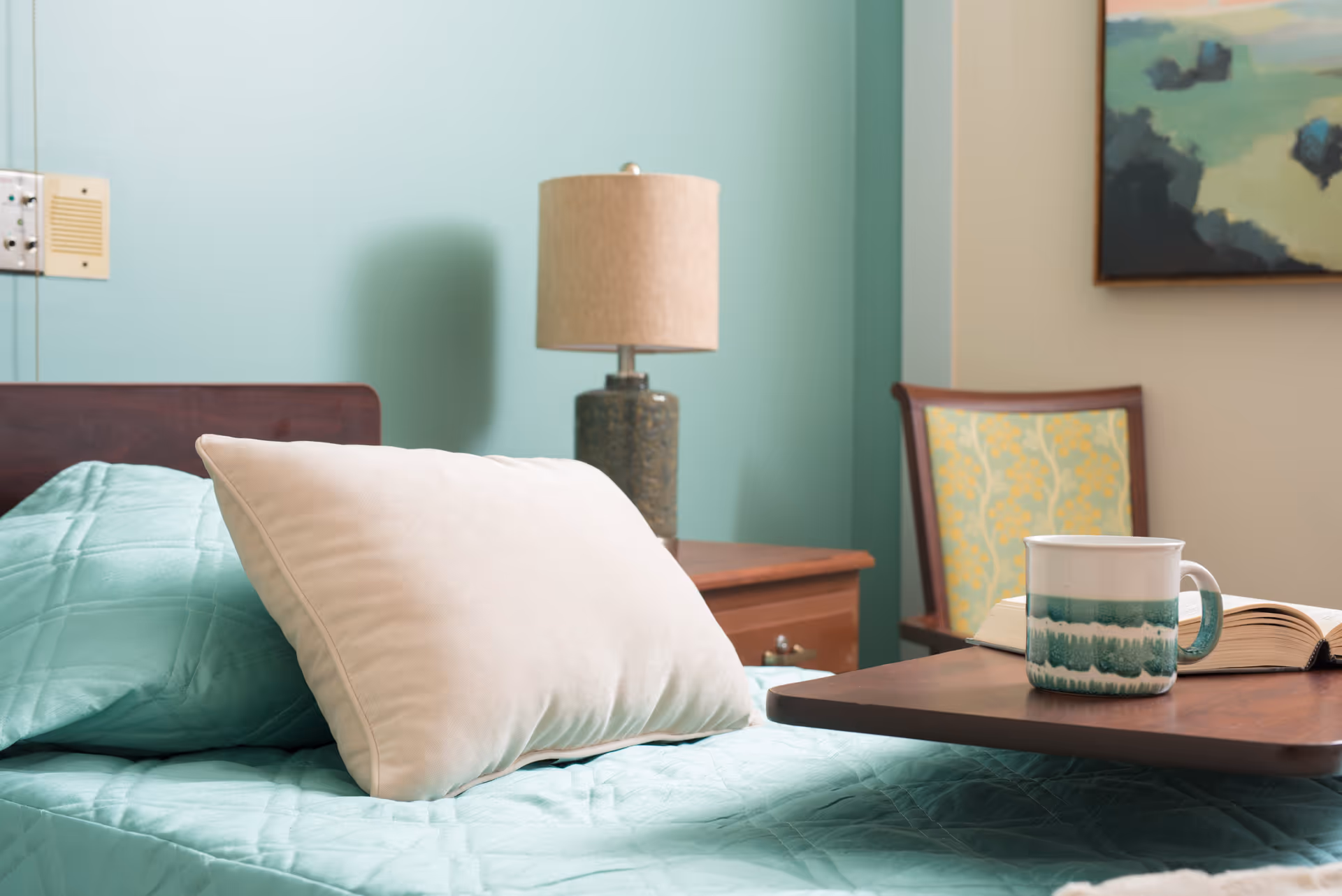 A neatly made bed with a light blue quilt and a large beige pillow in a room. Next to the bed is a wooden nightstand with a ceramic table lamp. In the background, there is a wooden chair with a patterned cushion and a painting on the wall. A wooden overbed table holds a ceramic mug and an open book.