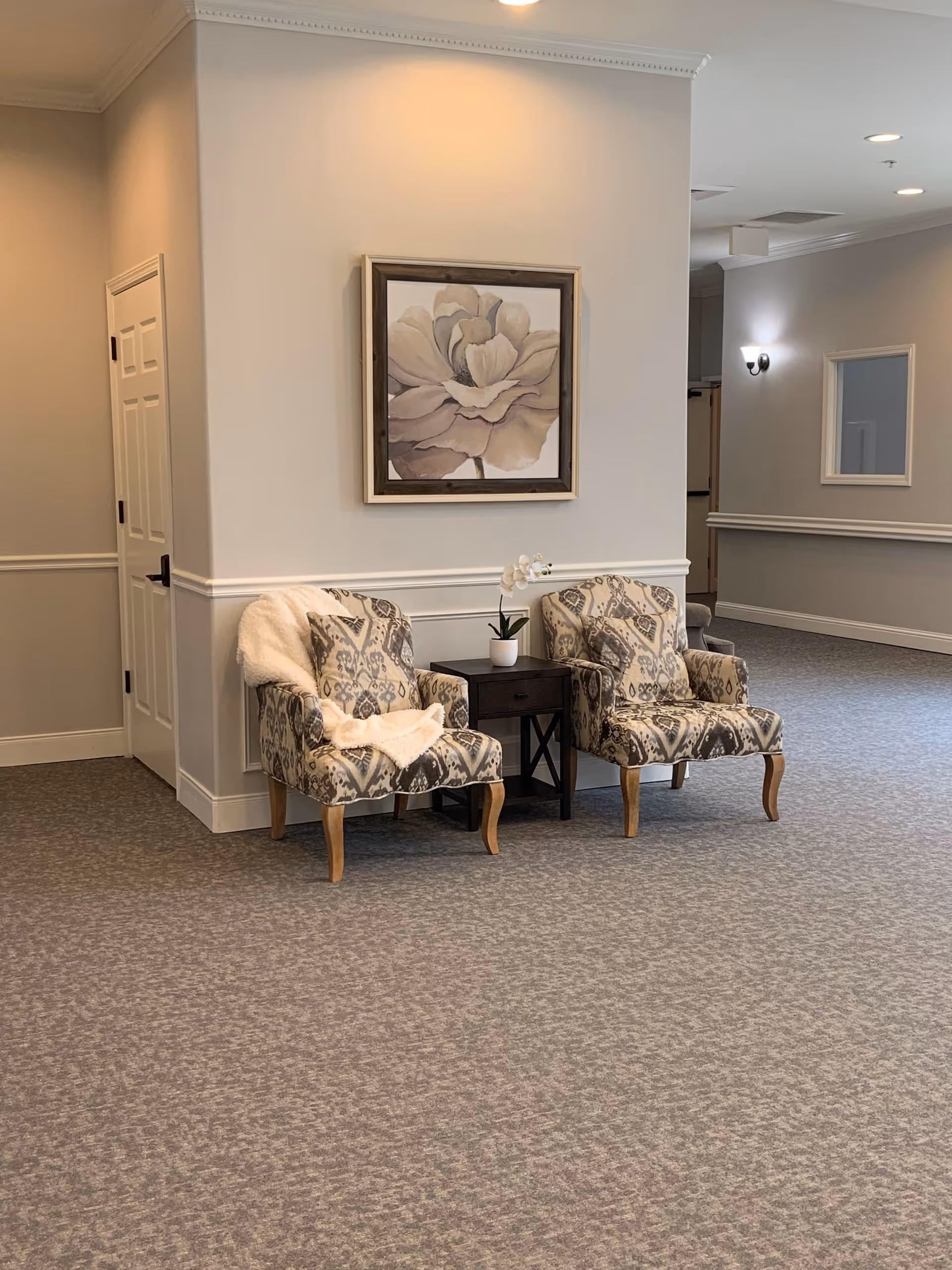 A cozy seating area in a hallway with two patterned armchairs separated by a small dark wooden table holding a white orchid plant. A soft white throw blanket is draped over one chair. Above the chairs hangs a framed painting of a large flower in neutral tones. The walls are painted light gray with white trim, and the floor is carpeted in a matching gray pattern. A white door is visible to the left, and a wall sconce and mirror are seen further down the hallway.