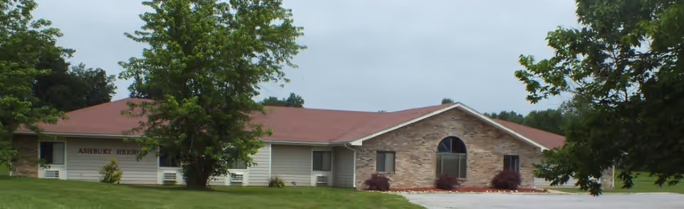 Single-story building with a red roof and beige siding surrounded by green grass and trees under a cloudy sky. The building has a sign that reads Ashbury Heights.