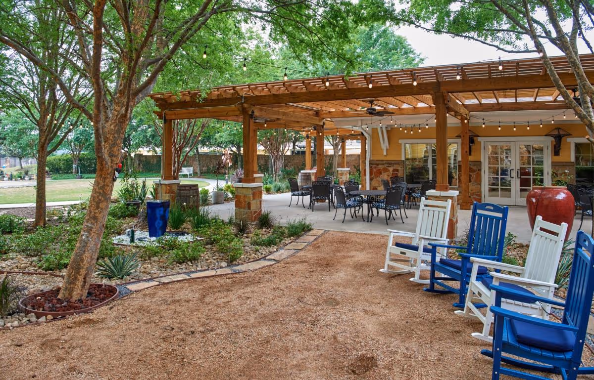 Outdoor seating area at The Village at Gleannloch Farms featuring a wooden pergola with string lights, several tables and chairs underneath, surrounded by trees and landscaped garden beds. There are also blue and white rocking chairs on a gravel pathway in the foreground.
