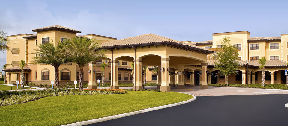 Exterior view of a large, multi-story senior living facility with a covered entrance, palm trees, well-maintained landscaping, and a paved driveway.