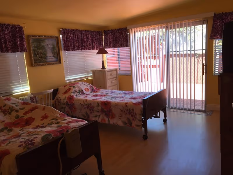 A senior living facility bedroom with two hospital-style beds covered in floral bedspreads. The room has yellow walls, wooden flooring, and windows with white blinds and purple valances. There is a white dresser with a lamp on top, a painting on the wall, and a sliding glass door with vertical blinds leading outside.