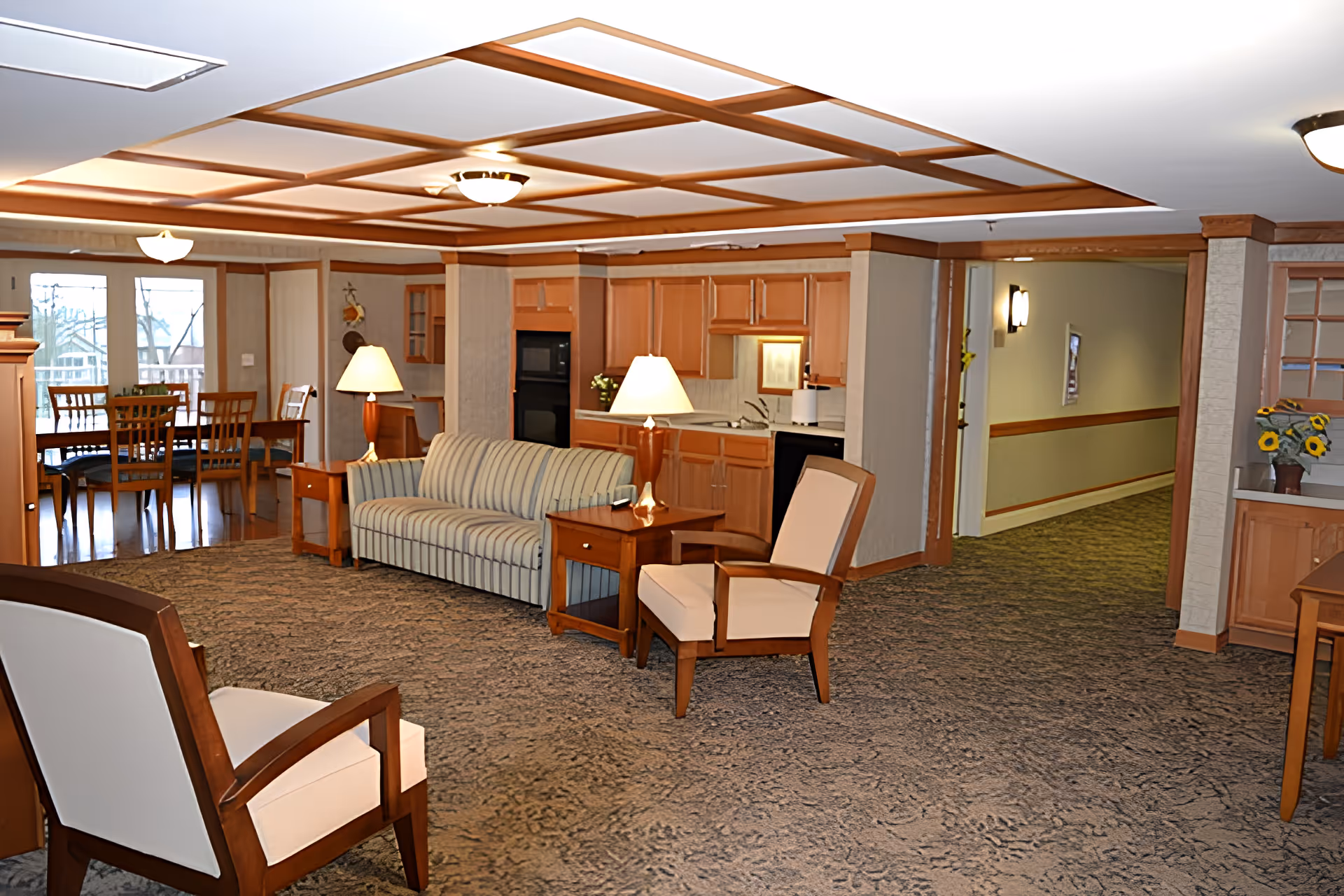 A spacious living room area in an assisted living facility featuring a striped sofa, two armchairs with wooden frames and cream cushions, two wooden side tables with lamps, and a carpeted floor. In the background, there is a dining area with a wooden table and chairs near large windows, and a kitchenette with wooden cabinets and appliances. The ceiling has a decorative wooden grid design with recessed lighting.