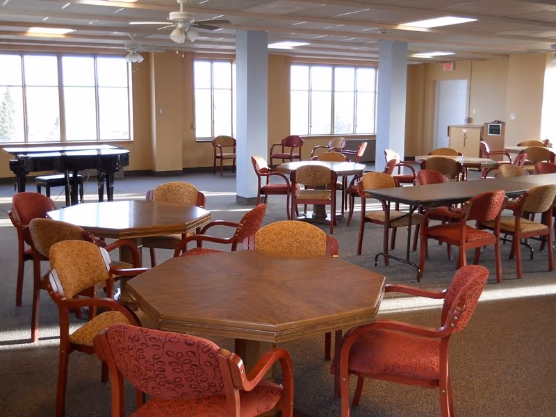 A spacious senior living dining hall with hexagonal tables, red chairs, a piano, and large windows.