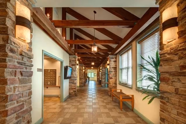 A long hallway in a senior living facility with stone pillars and wooden beams on the ceiling. The floor is tiled, and there are windows with blinds on the right side letting in natural light. A wooden bench and a potted plant are placed along the wall under the windows. Wall-mounted lights illuminate the corridor, and a flat-screen TV is mounted on the left wall near an open doorway.