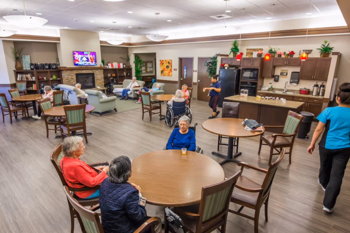 A spacious common area in a senior living facility with several elderly residents sitting at round wooden tables and in armchairs. The room features a kitchenette with cabinets, a refrigerator, and a microwave. There is a fireplace with a TV mounted above it, bookshelves, and plants decorating the space. Two staff members are walking through the room.