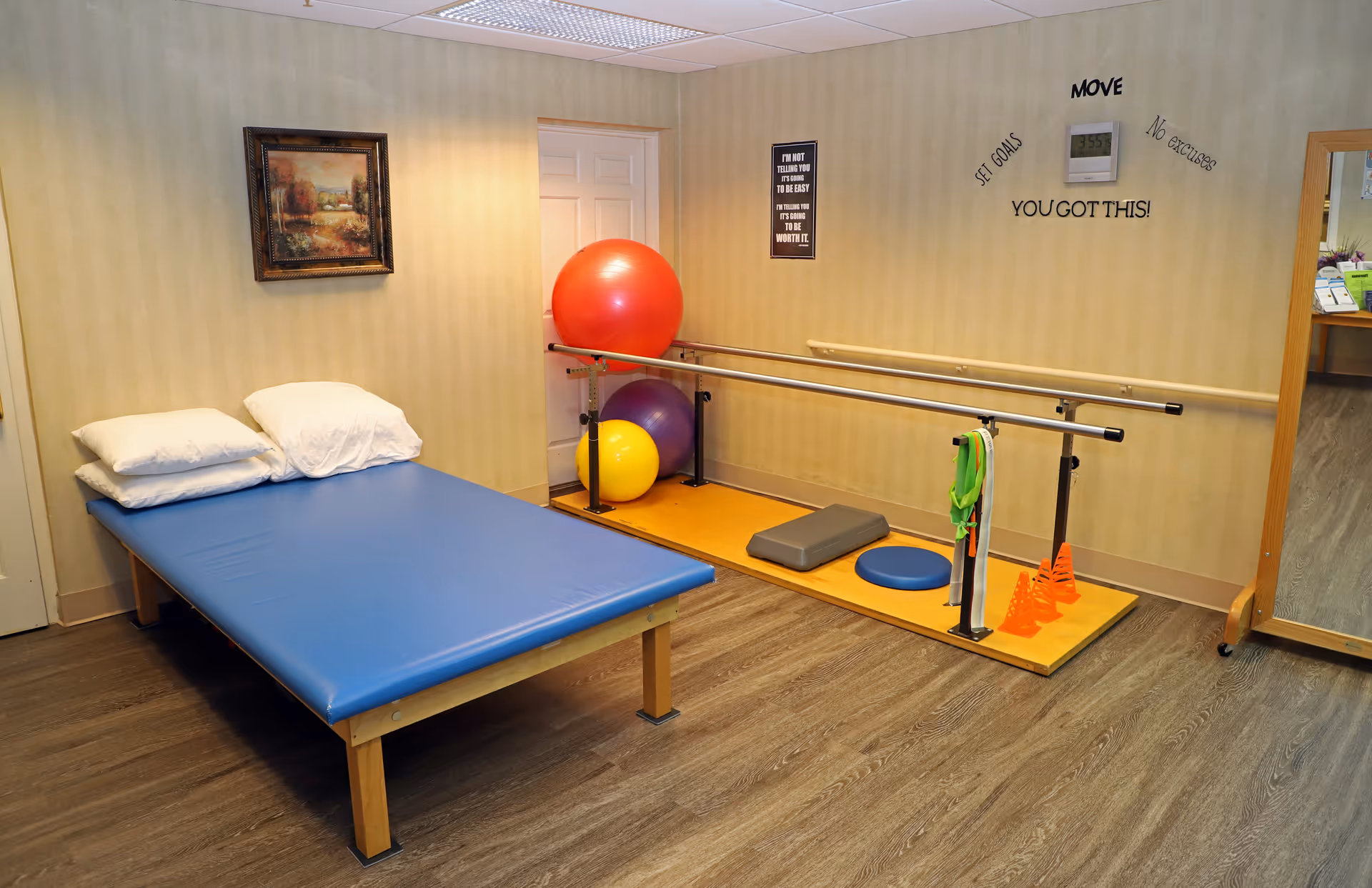 A physical therapy or rehabilitation room with a blue padded therapy table with pillows, parallel bars for walking exercises, colorful exercise balls, cones, and balance equipment. Motivational wall decals say 'MOVE', 'SET GOALS', 'No excuses', and 'YOU GOT THIS!'. A framed landscape painting hangs on the wall and a large mirror is visible on the right side.