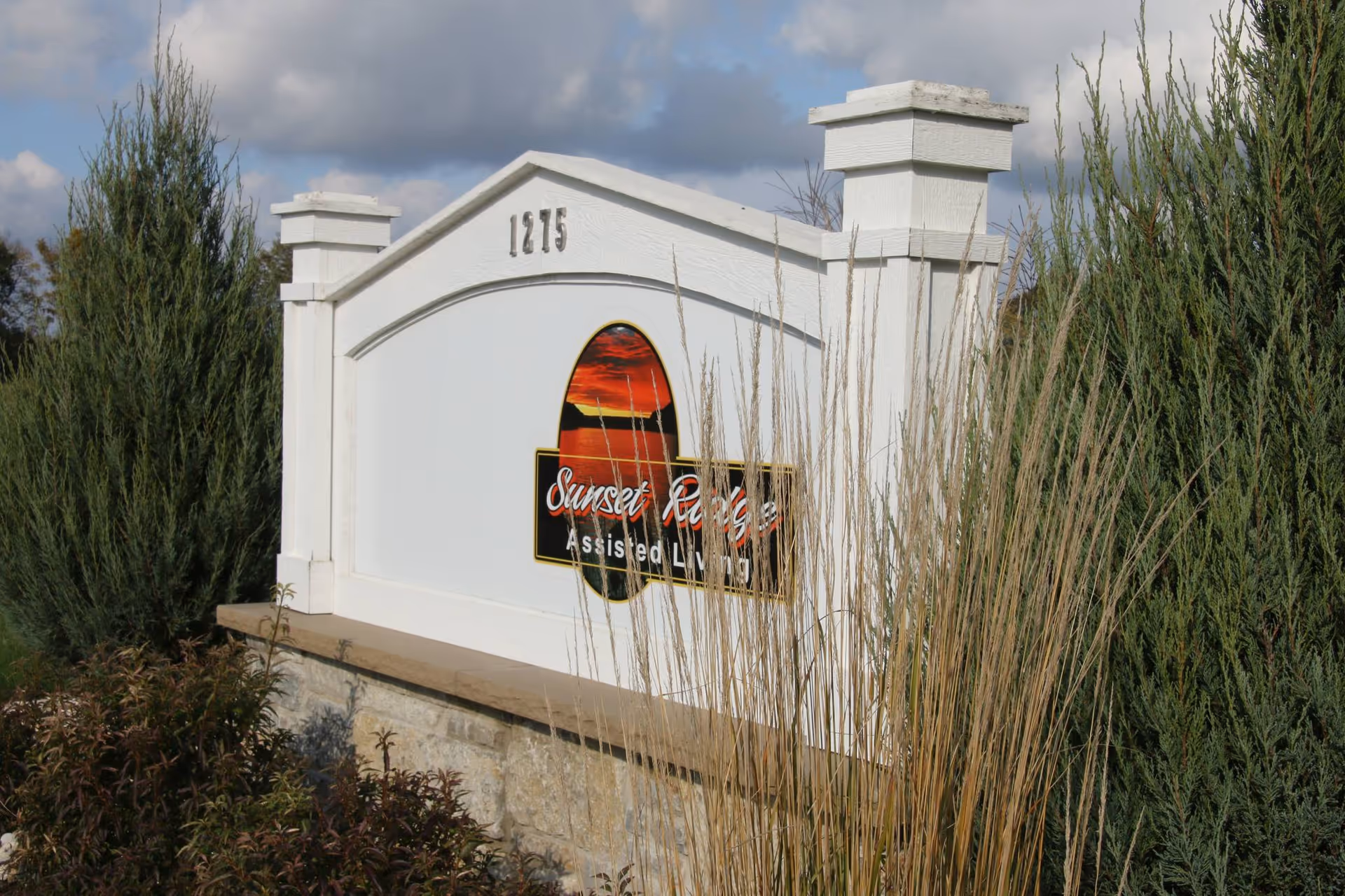 White stone and wood sign for Sunset Ridge Assisted Living with the number 1215 on top, surrounded by tall grasses and evergreen shrubs under a partly cloudy sky.