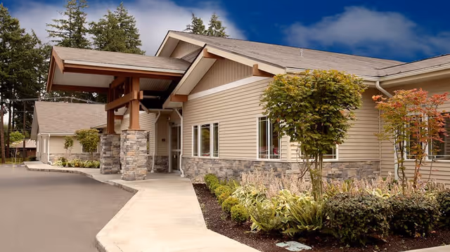 Exterior view of a single-story building with beige siding and stone accents, featuring a covered entrance supported by wooden beams. The building is surrounded by landscaped greenery and small trees under a partly cloudy blue sky.