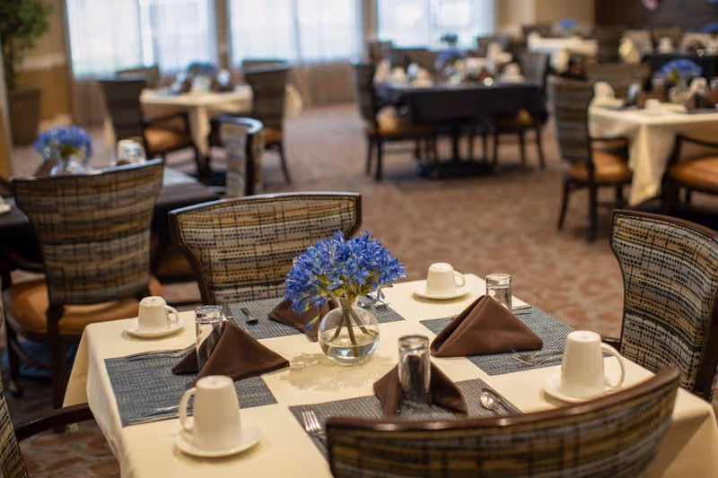 A dining room with tables set for four people, each place setting includes a white cup on a saucer, a glass, a brown folded napkin, and silverware. The tables are covered with cream-colored tablecloths and blue placemats. A small vase with blue flowers is centered on the table in the foreground. The room has multiple tables and chairs arranged neatly with soft lighting and large windows in the background.