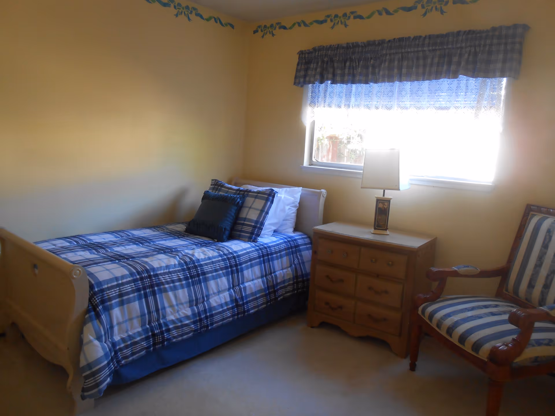 A small bedroom with a single bed covered in blue and white plaid bedding, a wooden nightstand with a lamp, a window with blue and white curtains, and a wooden armchair with blue and white striped upholstery.
