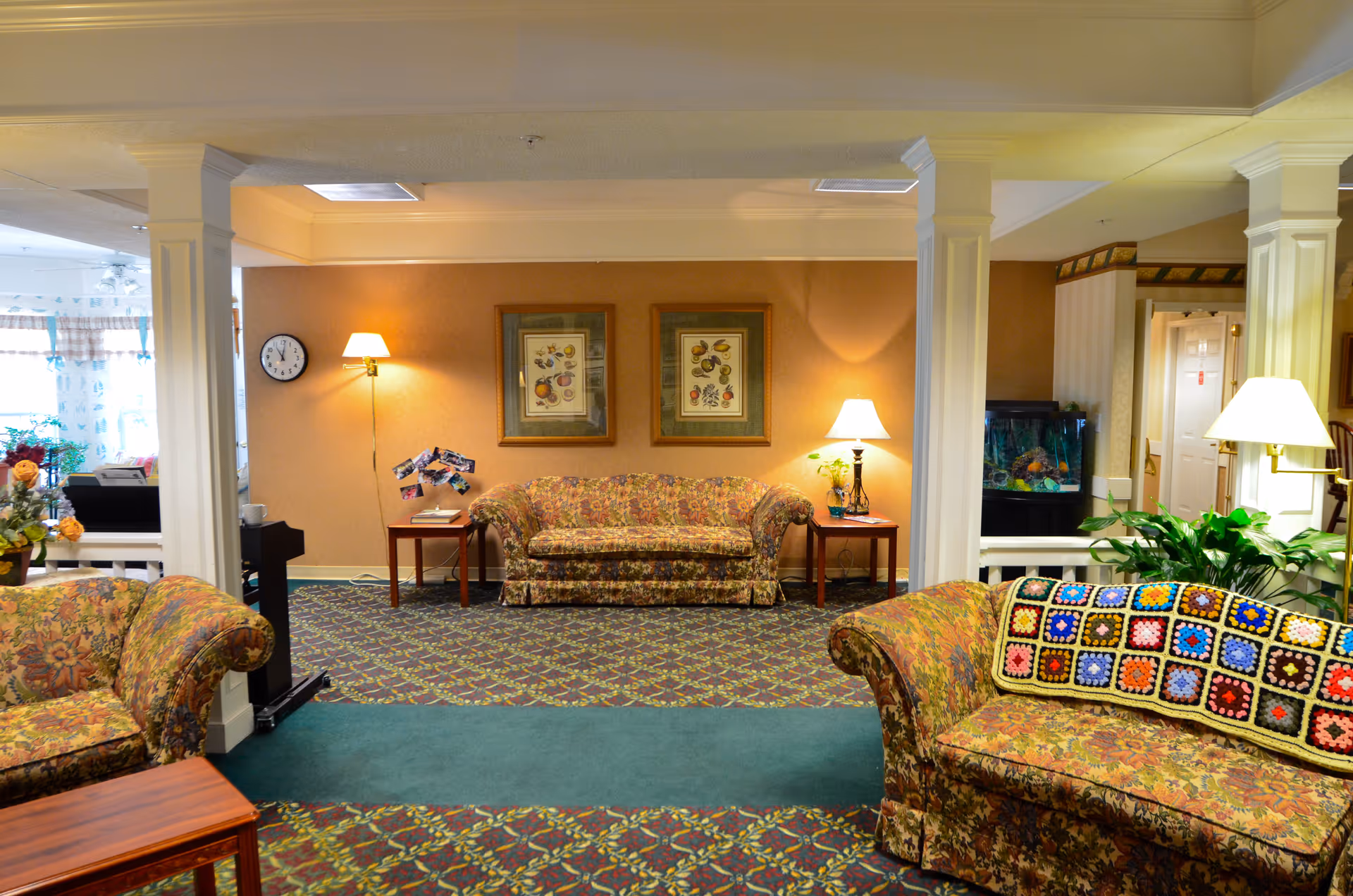A cozy living room area in a senior living facility with floral patterned sofas, two framed botanical prints on the wall, two side tables with lamps, a clock on the wall, and a fish tank in the background. The room has carpeted floors with a patterned design and white columns separating the space.