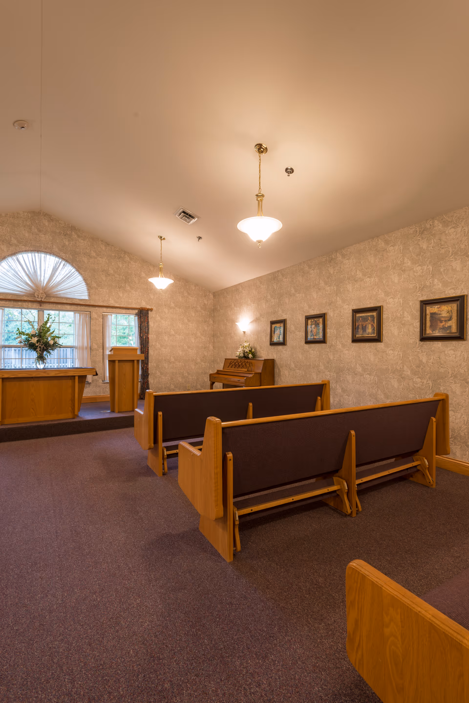 Interior of a small chapel or meditation room with wooden pews, a piano against the wall, framed pictures, and a podium near windows with a floral arrangement.