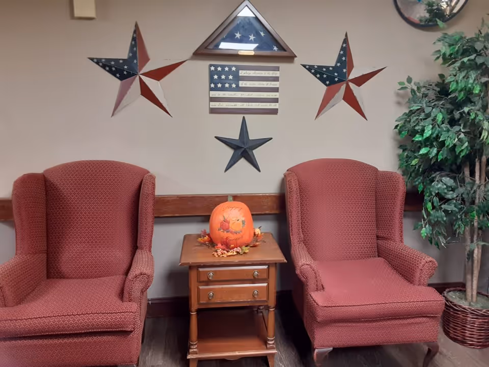 Two red upholstered armchairs with a small wooden side table between them, decorated with a pumpkin and autumn leaves. The wall behind features patriotic star decorations and a folded American flag in a triangular display case. A potted green plant is visible on the right side.