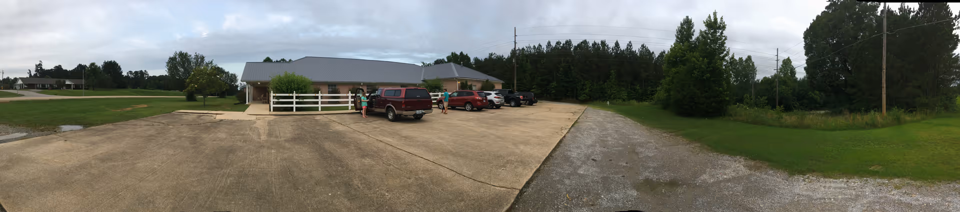 Panoramic view of a single-story assisted living building with a small parking lot of parked cars and surrounding grassy areas and trees under a cloudy sky.