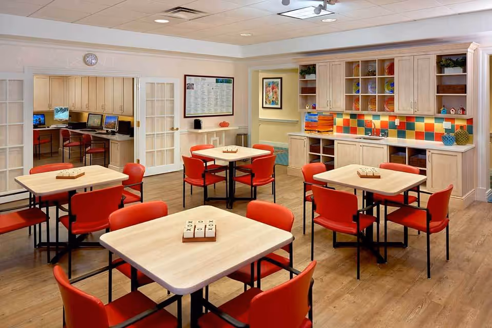 A bright and clean common area with four square tables, each surrounded by red chairs. On each table, there is a tic-tac-toe game set. The room features a colorful tiled backsplash above a countertop with cabinets and shelves holding decorative items. In the background, there is a small computer room with several computers and chairs visible through glass-paneled double doors. The floor is wooden, and the walls are light-colored with framed artwork.
