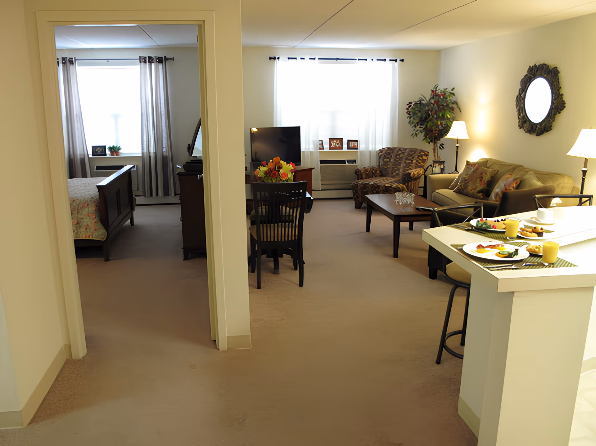 Furnished senior apartment interior showing a living room with sofa, armchair, TV and dining table, plus a bedroom visible through a doorway.