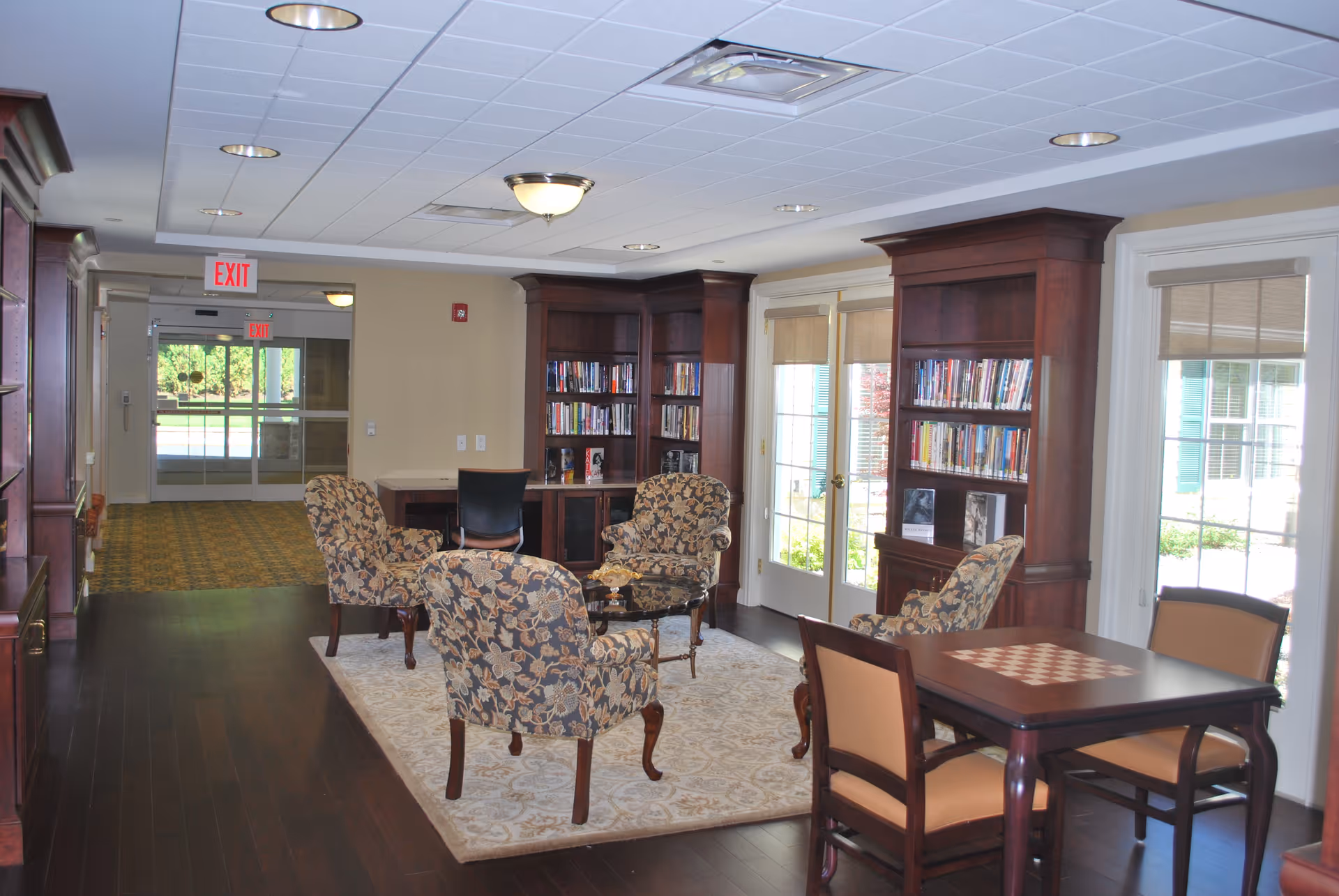 A cozy interior common area with floral upholstered armchairs arranged around a small glass coffee table on a patterned rug. There are two wooden bookshelves filled with books against the wall, and a table with a chessboard pattern on top with four chairs nearby. Large windows and glass doors let in natural light, and an exit sign is visible in the background.