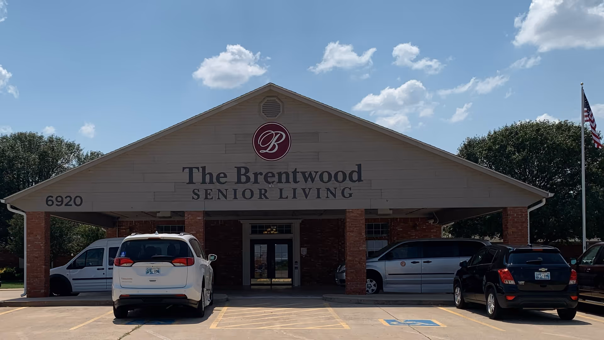 Front exterior view of The Brentwood Senior Living facility with a covered entrance supported by brick pillars. Several cars are parked in front, and an American flag is visible on the right side. The sky is partly cloudy.