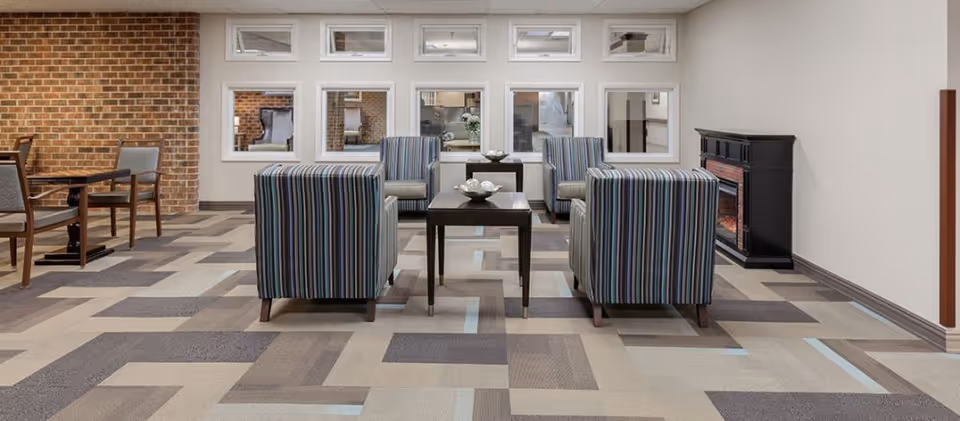 A modern supportive living common area with four striped armchairs arranged around two dark wooden coffee tables. The room features a patterned carpet in shades of beige, gray, and light blue, a brick accent wall on the left, and a black electric fireplace on the right. There are several windows on the far wall, allowing a view into an adjacent room.