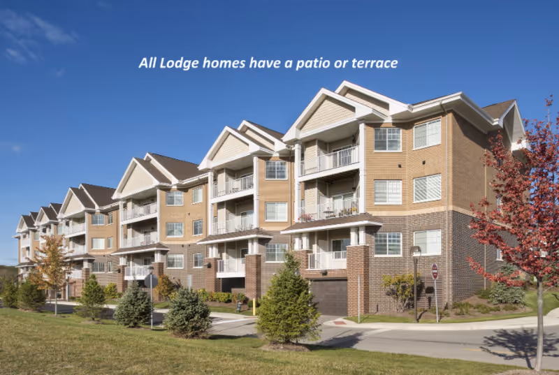 Exterior view of a multi-story senior living facility building with balconies and patios, surrounded by small trees and landscaping under a clear blue sky.